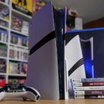PlayStation 5 Pro console displayed vertically with a DualSense controller positioned in front, shown on a shelf with video games in the background. This represents Sony's current generation while the company develops the significantly more powerful PS6.
