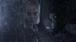 A rainy window with Grace Ashcroft's concerned face in close-up on the left side, while another female character can be seen in the background, both appearing distressed in what seems to be a dark, foreboding environment from Resident Evil Requiem.