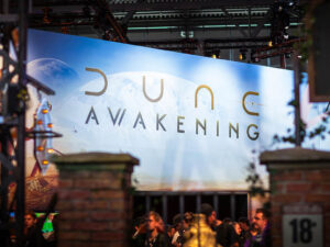 Dune Awakening logo displayed on a large blue-tinted screen at Gamescom 2025, showing the game title in gold and silver lettering against a backdrop featuring Arrakis with visible moons.